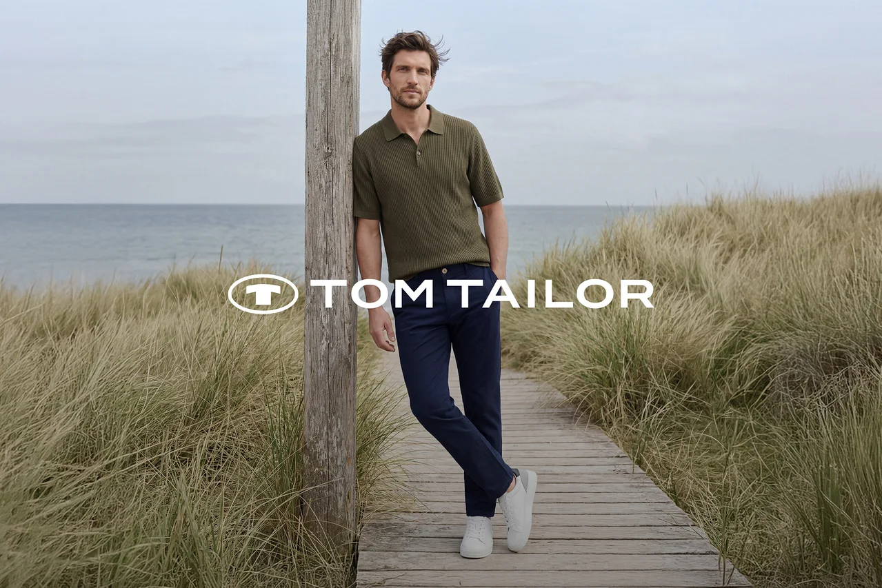 Muž v zelenom polo tričku, tmavomodrých nohaviciach a bielych teniskách sa opiera o drevený stĺp na drevenom chodníku pri mori, obklopený vysokou trávou. Logo TOM TAILOR.