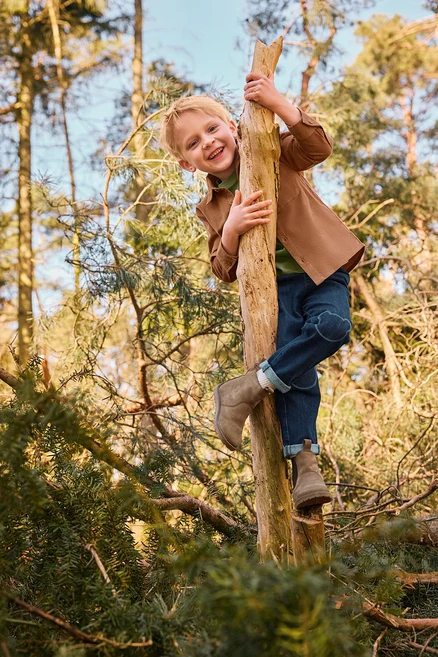 Usmievavý chlapec lezie na kmeň stromu v lese, oblečený v hnedej bunde, zelenom tričku, modrých džínsoch s nášivkami na kolenách a svetlohnedých čižmách.