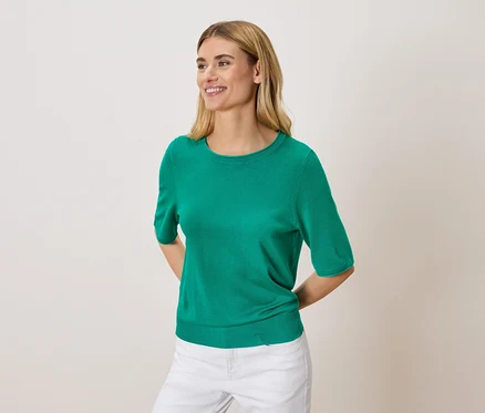 Žena s blond vlasmi má na sebe zelený sveter a biele nohavice.