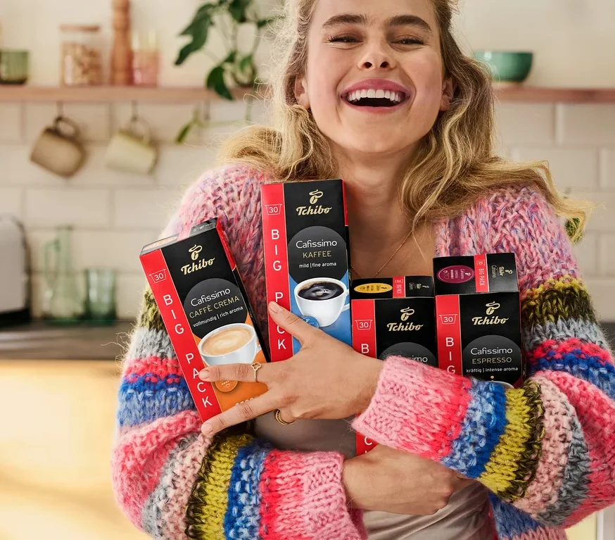 Usmievajúca sa žena drží balíčky kávových kapsúl Tchibo Cafissimo v kuchyni.