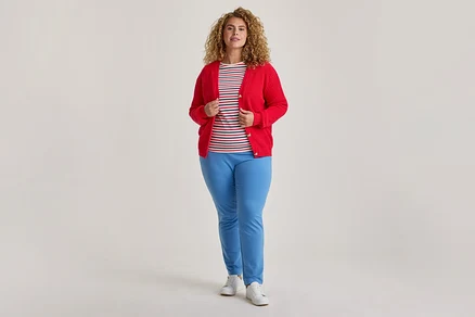 Žena s kučeravými vlasmi stojí, oblečená v rozopnutom červenom kardigáne, pruhovanom tričku, svetlomodrých nohaviciach a bielych teniskách.