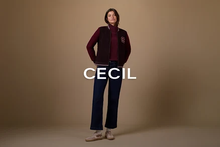 Žena stojí s rukami vo vreckách, oblečená v tmavočervenom roláku, tmavohnedej bomber bunde s bielym nášivkou „B“, tmavomodrých rozšírených džínsoch a svetloružových teniskách s bielymi ponožkami, na jednofarebnom hnedom pozadí.