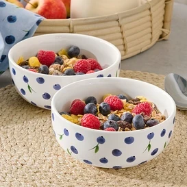 Dve misky s müsli, malinami a čučoriedkami na prestieraní. V pozadí džbán s mliekom a jablká v košíku.
