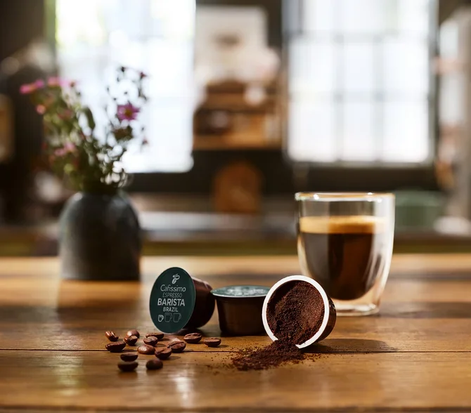 Na drevenom stole stojí pohár s espressom, vedľa neho sú tri kapsuly Espresso Brasil a rozsypaná zrnková káva.