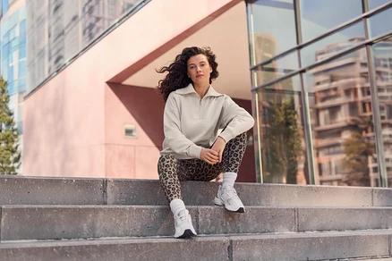Žena s kučeravými vlasmi sedí na betónových schodoch, oblečená v béžovom tope s polovičným zipsom, legínach s leopardím vzorom a bielych teniskách, s modernými budovami v pozadí.