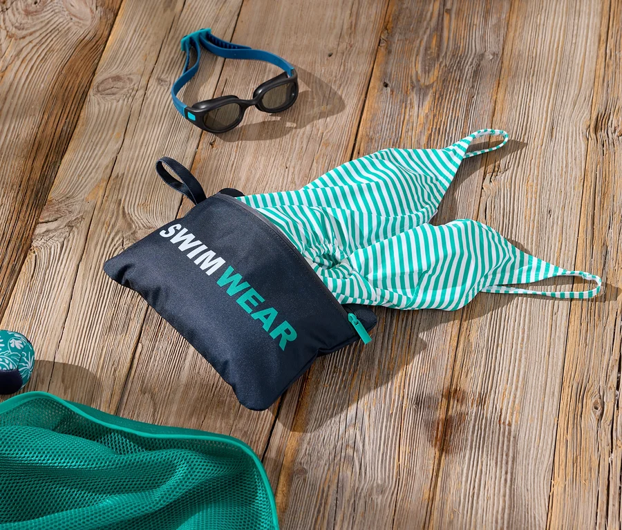 Snímek plaveckých doplňků na dřevěném povrchu včetně tašky s nápisem „Swimwear“, zeleno-bílých pruhovaných plavek, plaveckých brýlí a zelené síťky.