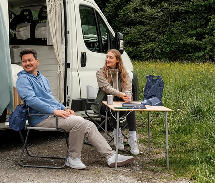 Muž v svetlomodrej teplákovej mikine s kapucňou a žena v béžovej teplákovej bunde a kaki hybridnej veste sedia pred karavanom. Na stole leží tmavomodrá zbaliteľná vesta.