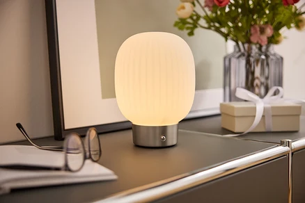 Nočná lampa s bielym tienidlom stojí na skrinke vedľa okuliarov, knihy, vázy s kvetmi a darčekovej krabičky.