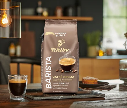 Balenie BARISTA Caffè Crema - 1× 1 kg zrnková káva stojí na stole vedľa pohára kávy.