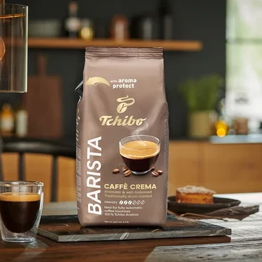 Balenie BARISTA Caffè Crema - 1× 1 kg zrnková káva stojí na stole vedľa pohára kávy.
