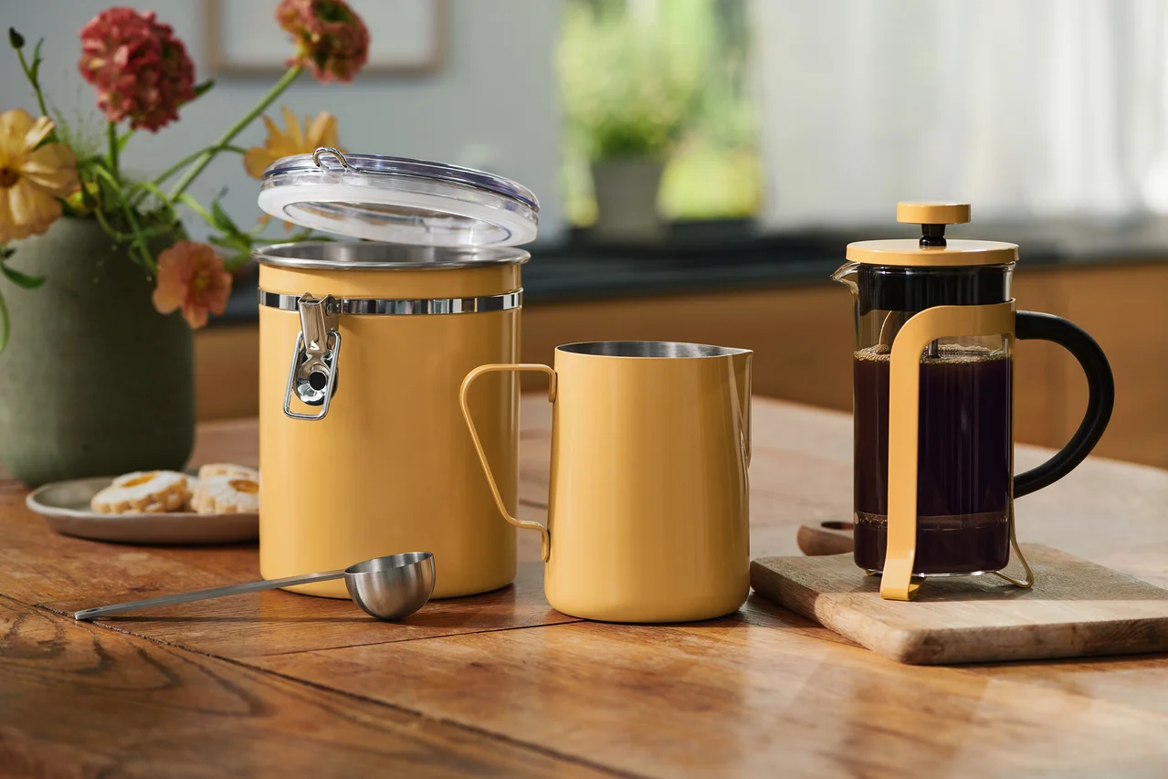 Žltá úložná nádoba s otvoreným vekom, zodpovedajúci hrnček, odmerka a French Press s kávou na drevenom stole. Kvety a sušienky v pozadí.