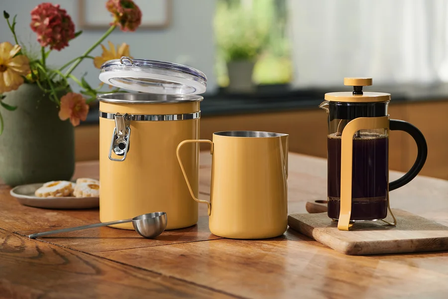 Žltá úložná nádoba s otvoreným vekom, zodpovedajúci hrnček, odmerka a French Press s kávou na drevenom stole. Kvety a sušienky v pozadí.