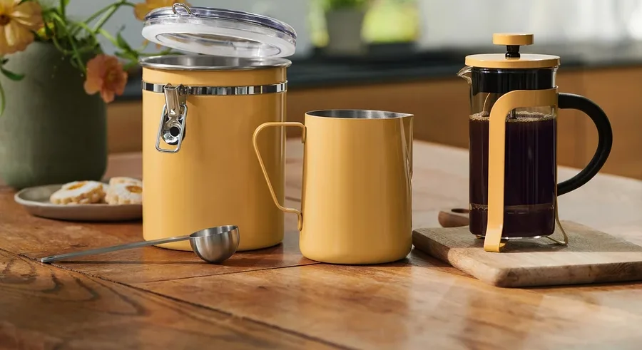 Žltá úložná nádoba s otvoreným vekom, zodpovedajúci hrnček, odmerka a French Press s kávou na drevenom stole. Kvety a sušienky v pozadí.
