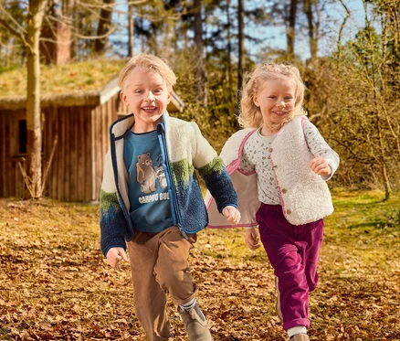 Dve usmievavé deti, chlapec a dievča, bežia cez listnatú vonkajšiu oblasť. Chlapec má na sebe modré tričko s grafikou, farebnú flísovú bundu a hnedé menčestrové nohavice. Dievča má na sebe vzorované tričko, krémovú chlpatú vestu a purpurové menčestrové nohavice. V pozadí je drevený dom s trávnatou strechou.