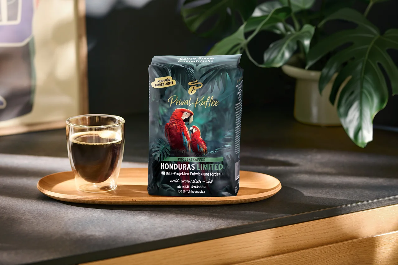 Tmavozelené balenie kávy „Privat Kaffee Honduras Limited“ s dvoma červenými arami a sklenená šálka čiernej kávy stoja na drevenom podnose. Na balení sú nápisy ako „NUR FÜR KURZE ZEIT!“ a „100% Tchibo Arabica“. V pozadí je zelená rastlina.
