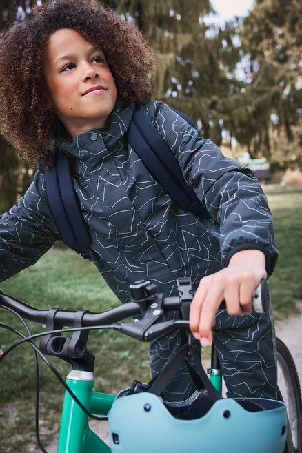 Dieťa s kučeravými vlasmi v tmavej vzorovanej bunde a ruksaku stojí pri zelenom bicykli, svetlomodrá prilba visí na riadidlách, pozerá sa hore.