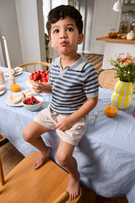 Chlapec sedí na okraji stola s jahodou na vidličke, má na sebe Kinder-Shorts a Kinder-Strick-Poloshirt. Na stole váza a podnos na tortu.