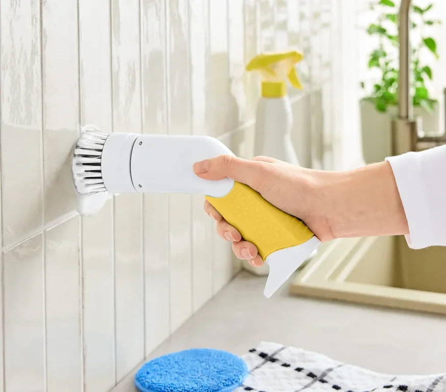Ruka drží elektrickú čistiacu kefu so žltou rukoväťou a bielou hlavicou, čistiacu dlaždice.