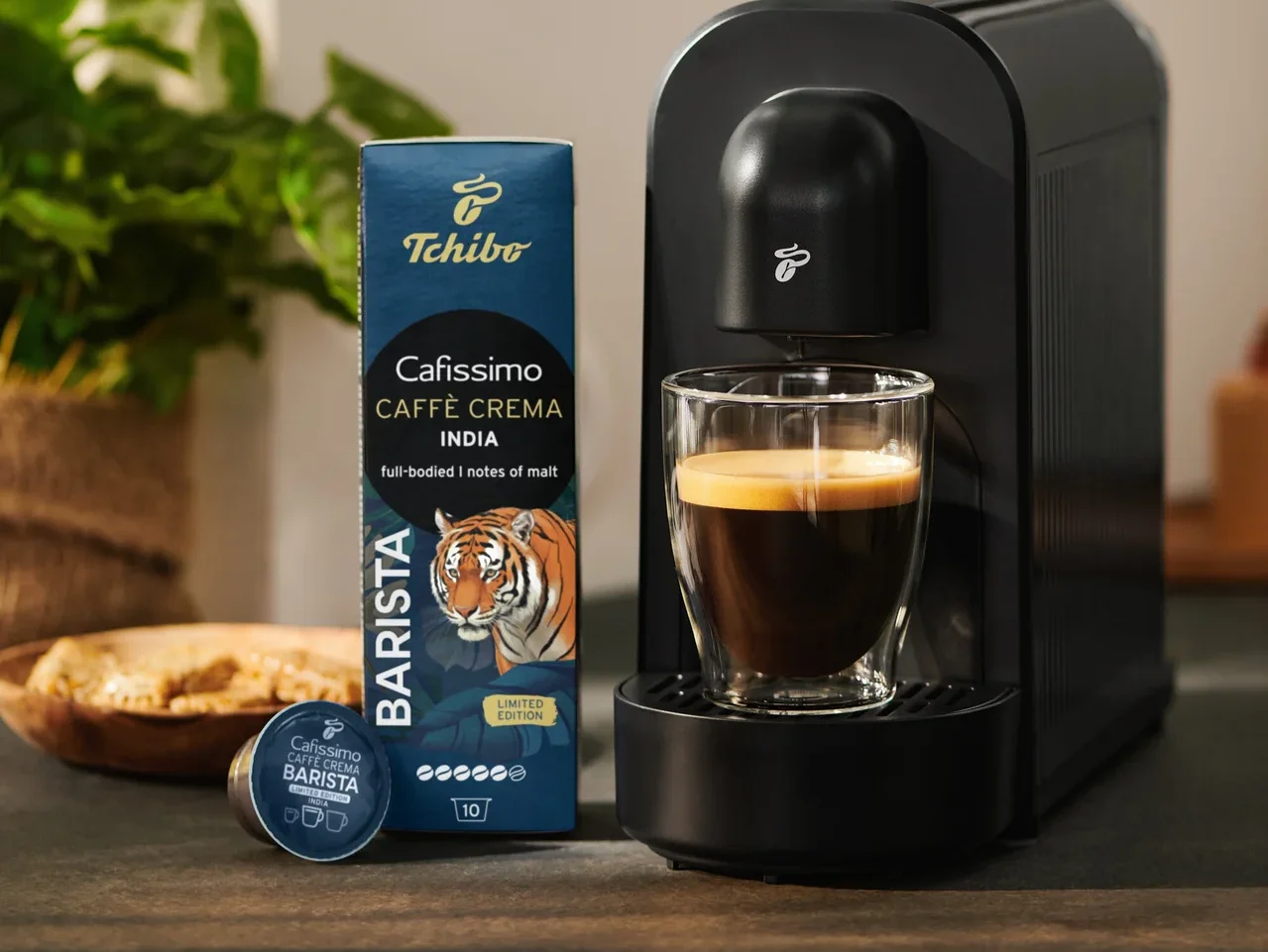 Čierny kávovar varí kávu do skleneného pohára, vedľa neho je modré balenie kapsúl Tchibo Cafissimo Caffè Crema India a jedna kapsula.