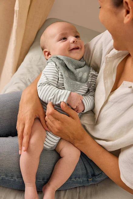 Matka drží usmievajúce sa dieťa, ktoré má na sebe body s možnosťou prispôsobenia veľkosti a mušelínovú šatku.