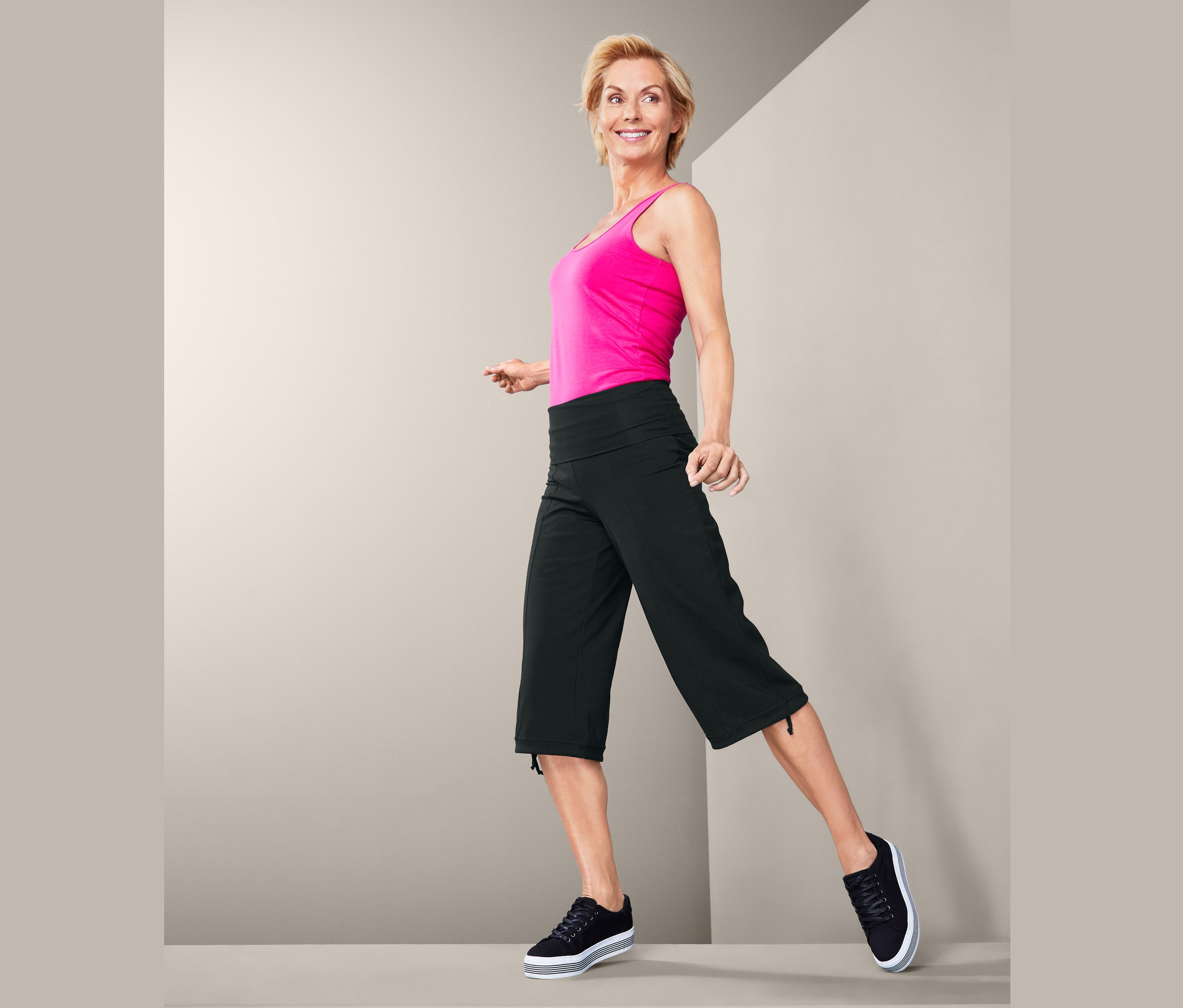 Žena v ružovom tielku a trojštvrťových športových nohaviciach stojí na sivom pozadí.