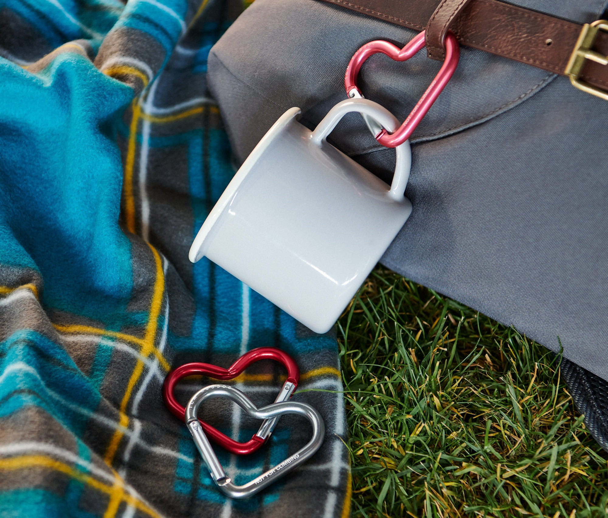 Na tráve leží deka a batoh, ku ktorému je pomocou karabíny v tvare srdca pripevnený hrnček. Dve karabíny v tvare srdca ležia na deke.