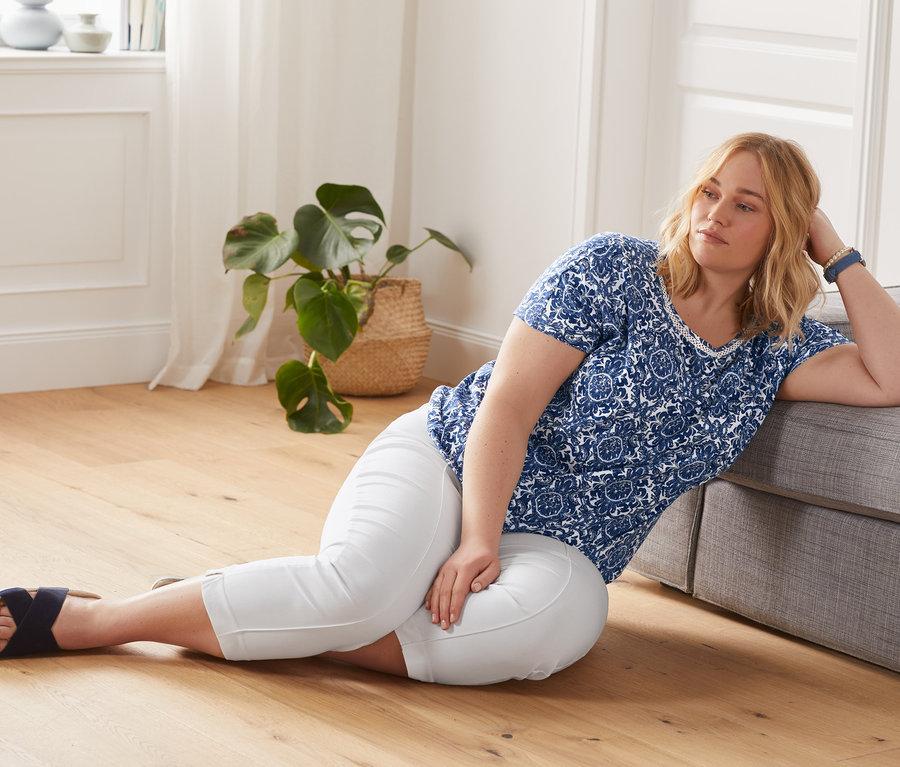 Žena leží na podlahe a opiera sa o pohovku. Má na sebe biele sedemosminové elastické nohavice a modré blúzkové tričko s potlačou.