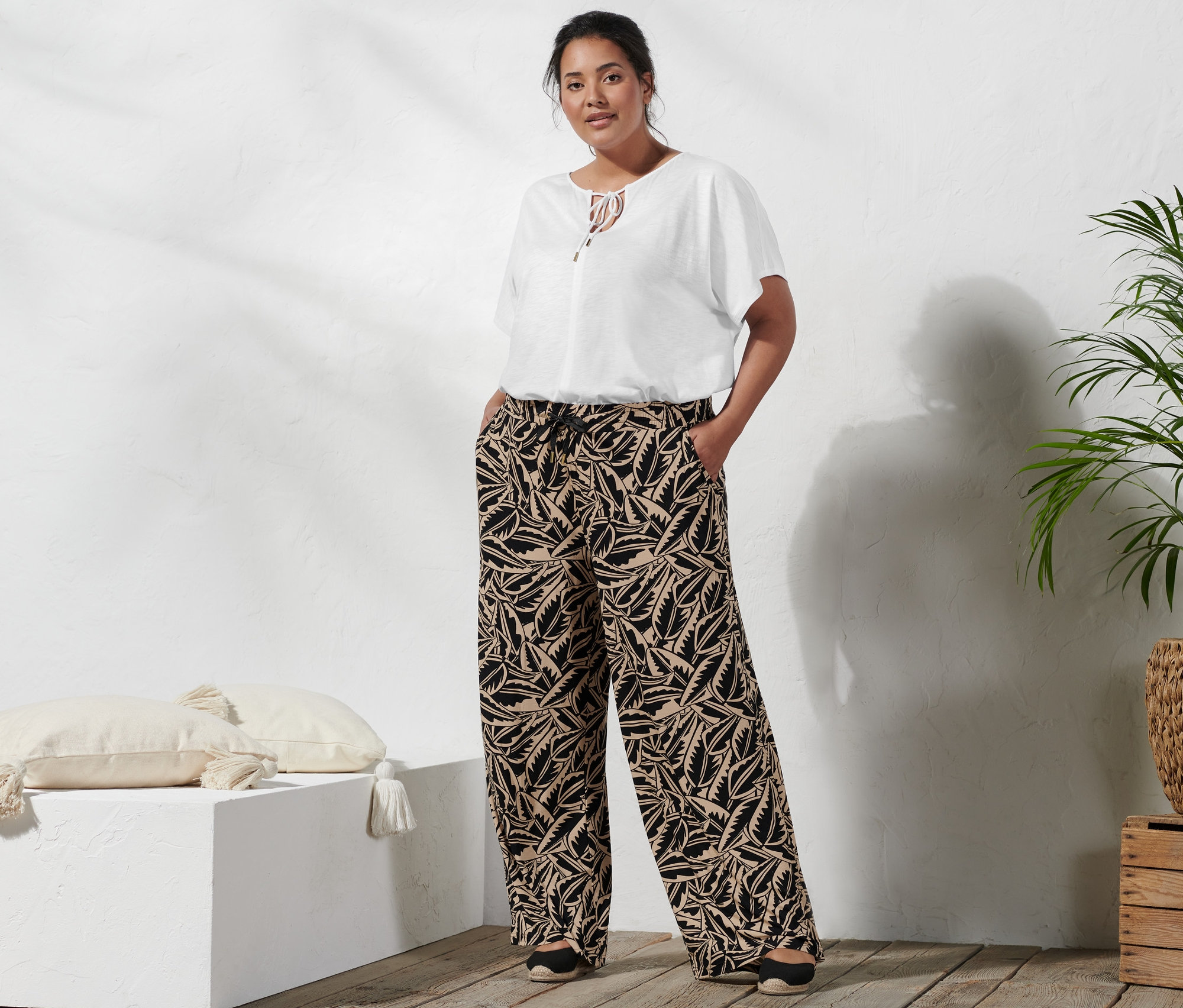 Žena stojí a má ruky vo vreckách nohavíc Palazzo s potlačou, kombinácia čiernej a béžovej, na sebe má blúzkové tričko so šnúrkou na zaväzovanie a espadrilky s klinovým podpätkom.