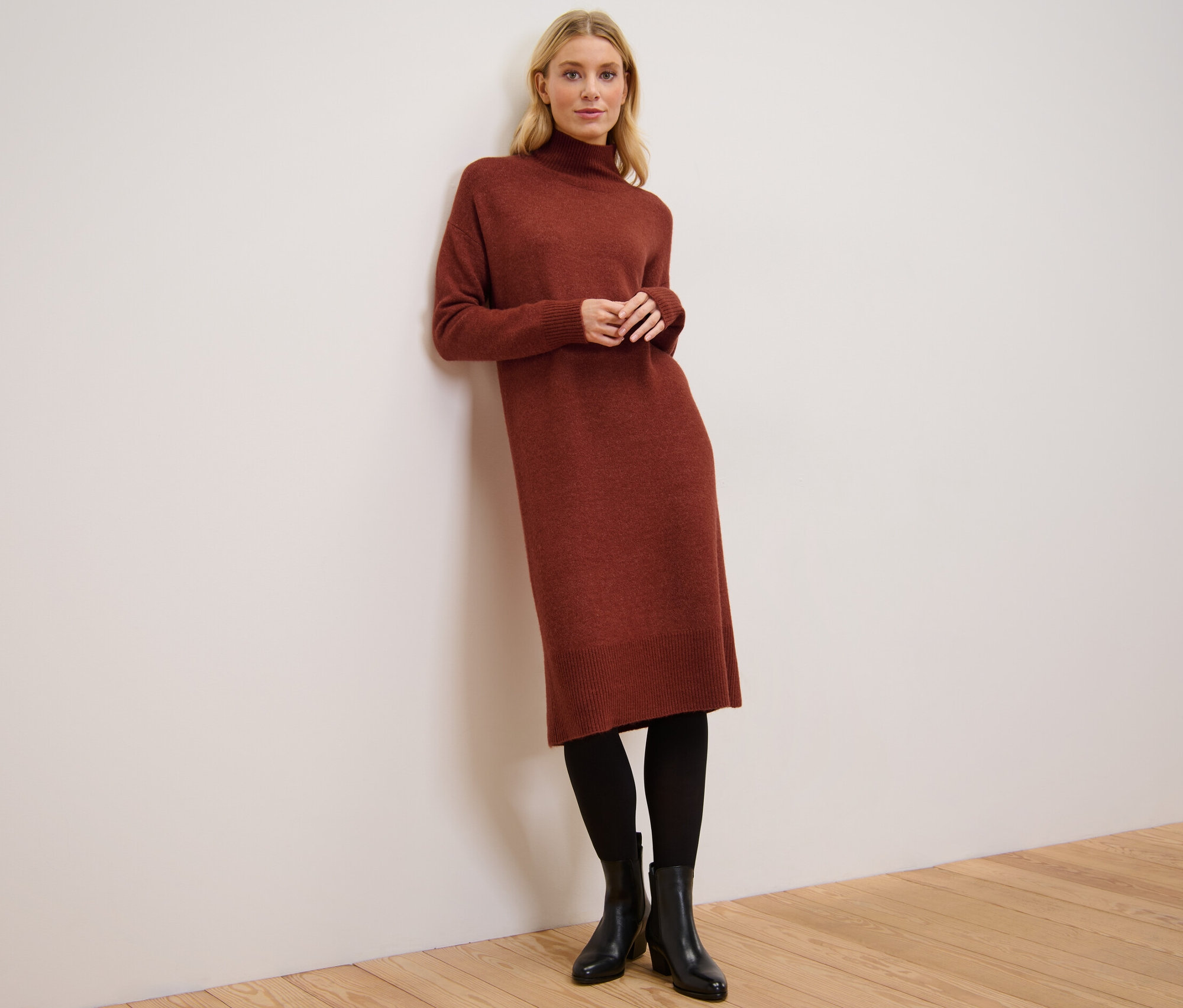 Žena opretá o stenu má na sebe pletené šaty s podielom vlny, čierne pančuchy a topánky.
