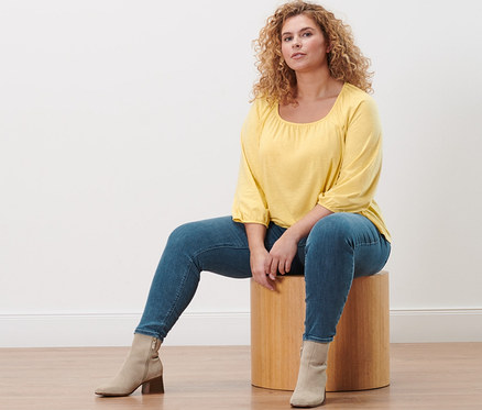 Žena sedí na drevenej stoličke a má na sebe žlté blúzkové tričko s 3/4 rukávom a tregínsy »Fit Nelly«.