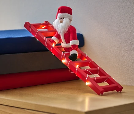 Bábika Santa Claus lezie po červenom rebríku s LED svetelnou reťazou, ktorý je opretý o hromadu kníh.