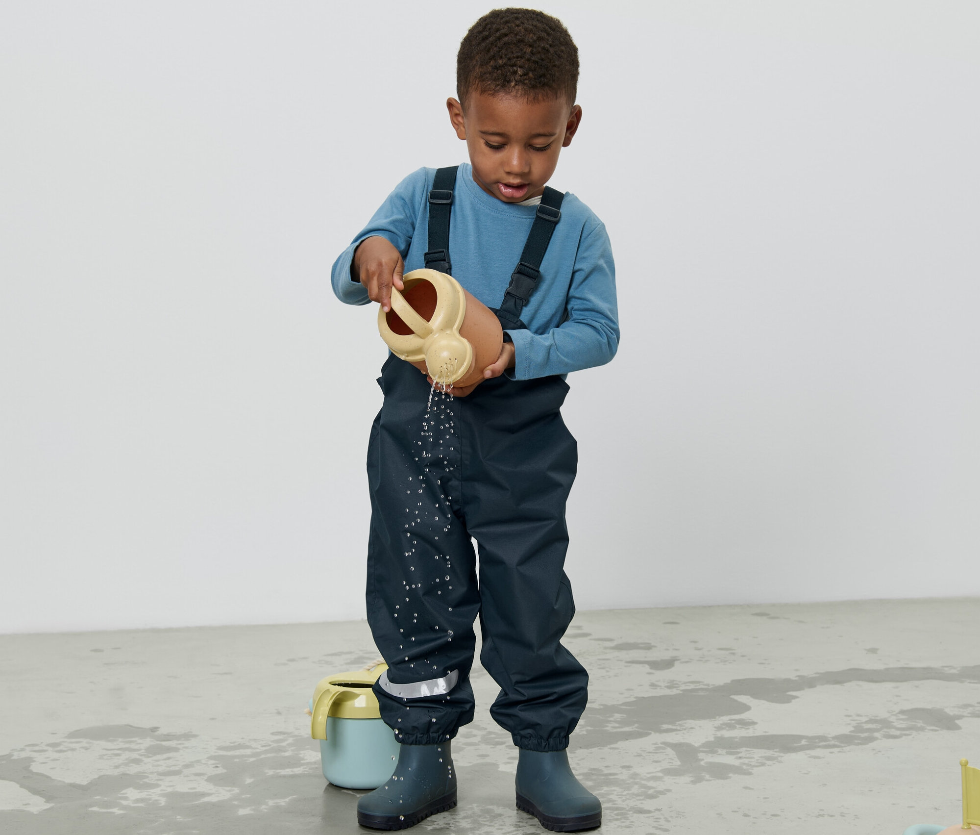 Chlapec v detských nohaviciach do dažďa leje vodu z kanvy na zem.