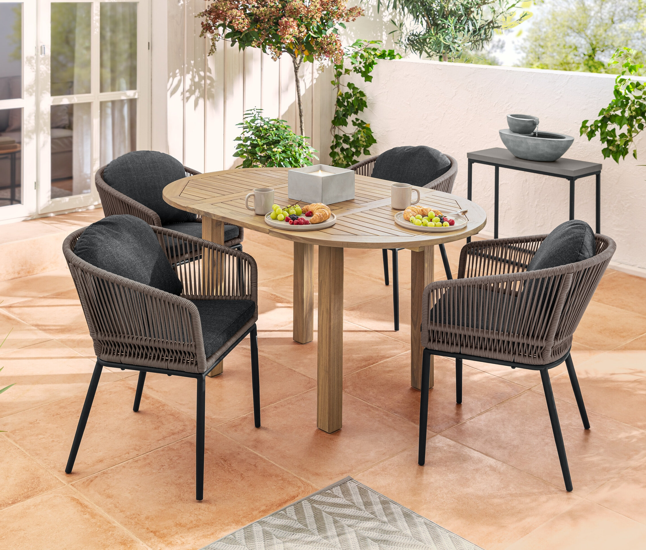 Štyri jedálenské kreslá s okrúhlou opierkou a textilným výpletom okolo okrúhleho rozkladacieho stola s jedlom a pitím na terase. V pozadí solárna záhradná fontána a súprava konzolových stolíkov.