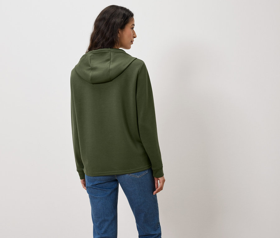 Žena chrbtom k fotoaparátu má na sebe zelenú mikinu s kapucňou a džínsy.