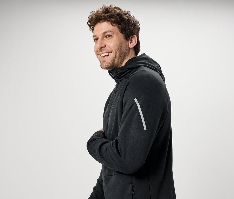 Usmievajúci sa muž s kučeravými vlasmi má na sebe čiernu mikinu s kapucňou.