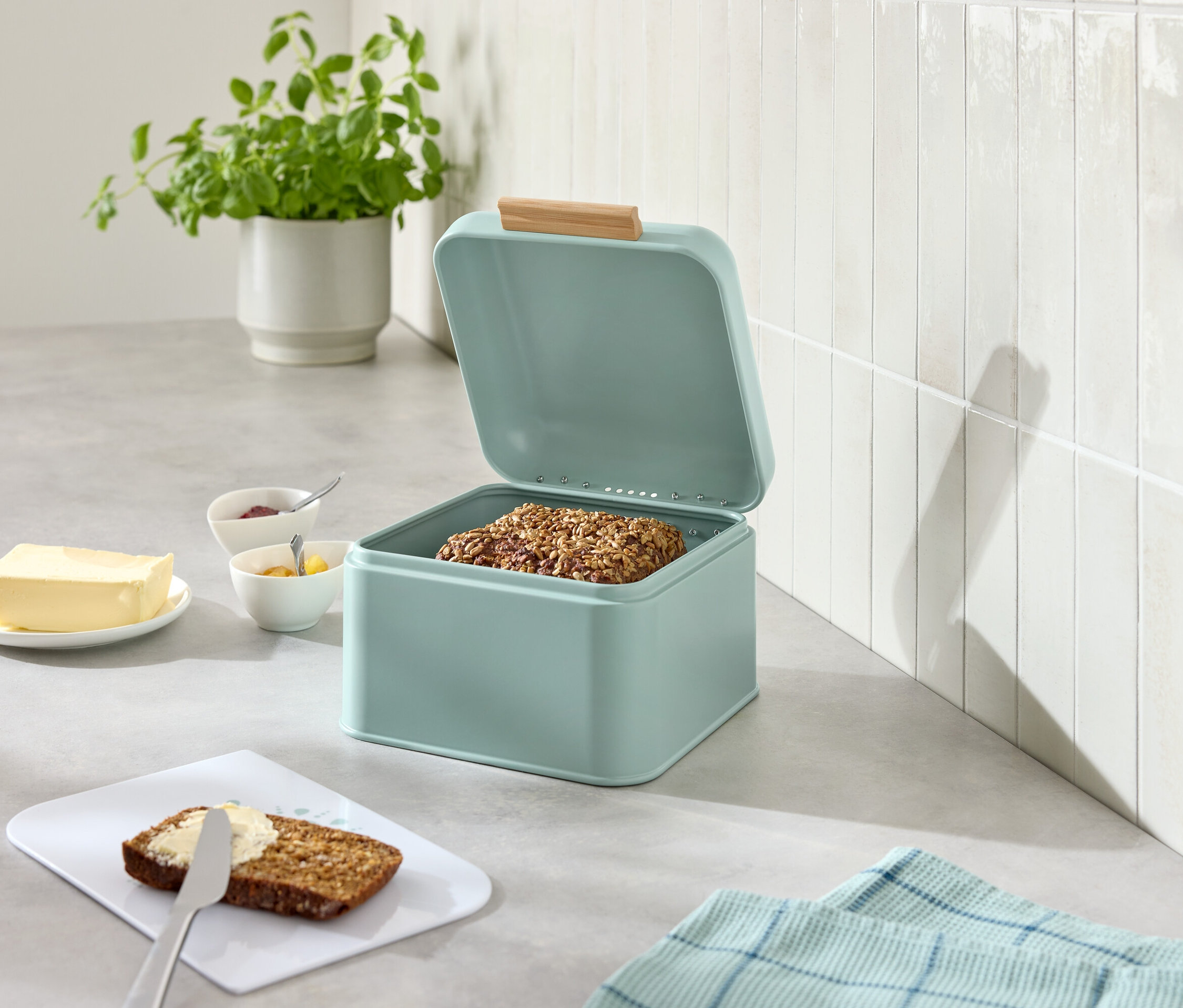 Otvorená svetlomodrá chlebovka s chlebom, vedľa maslo, džem, nôž a natretý chlieb.