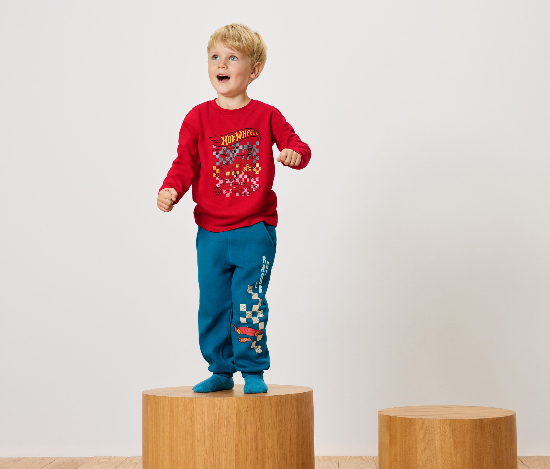 Chlapec stojí na drevenom podstavci v červenom tričku a modrých nohaviciach.