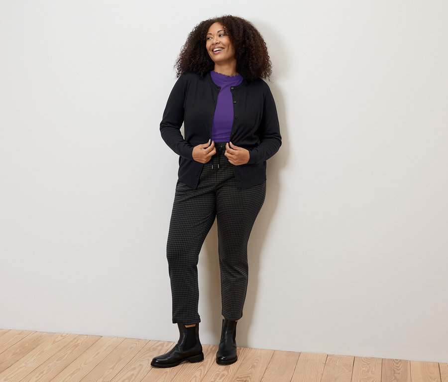 Žena má na sebe fialové tričko s vlnitým lemom, tmavé nohavice a sveter.