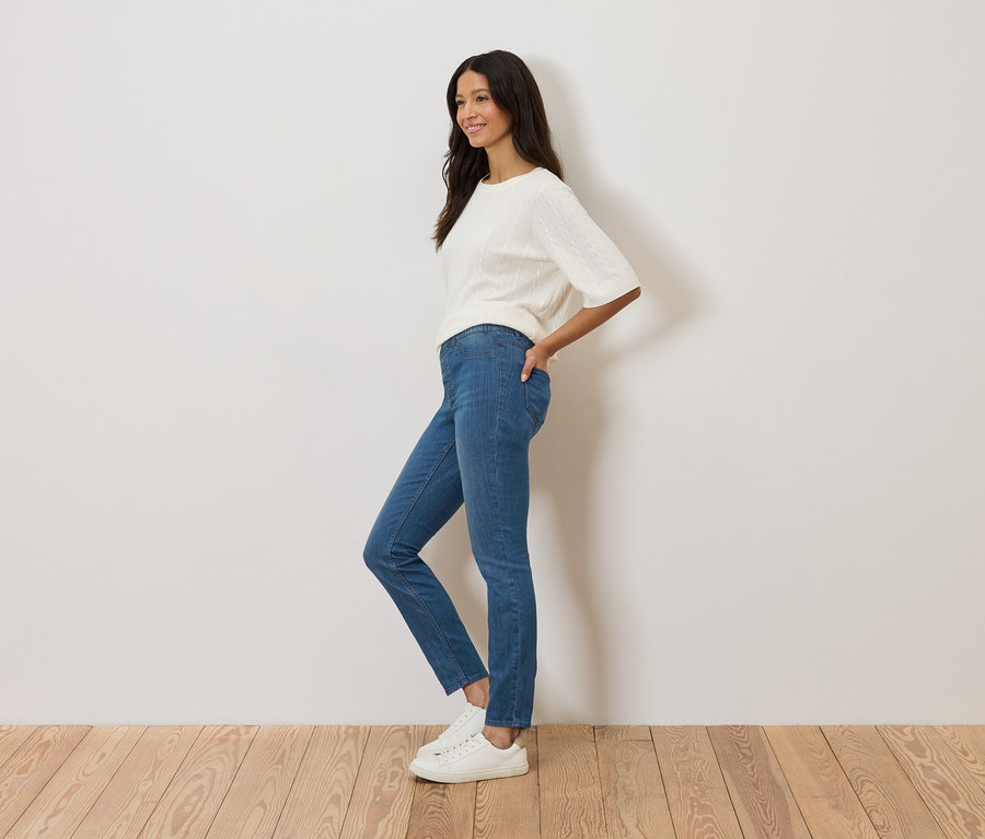 Žena stojí bokom pred bielou stenou, má na sebe modré džínsy, biely sveter a biele tenisky.