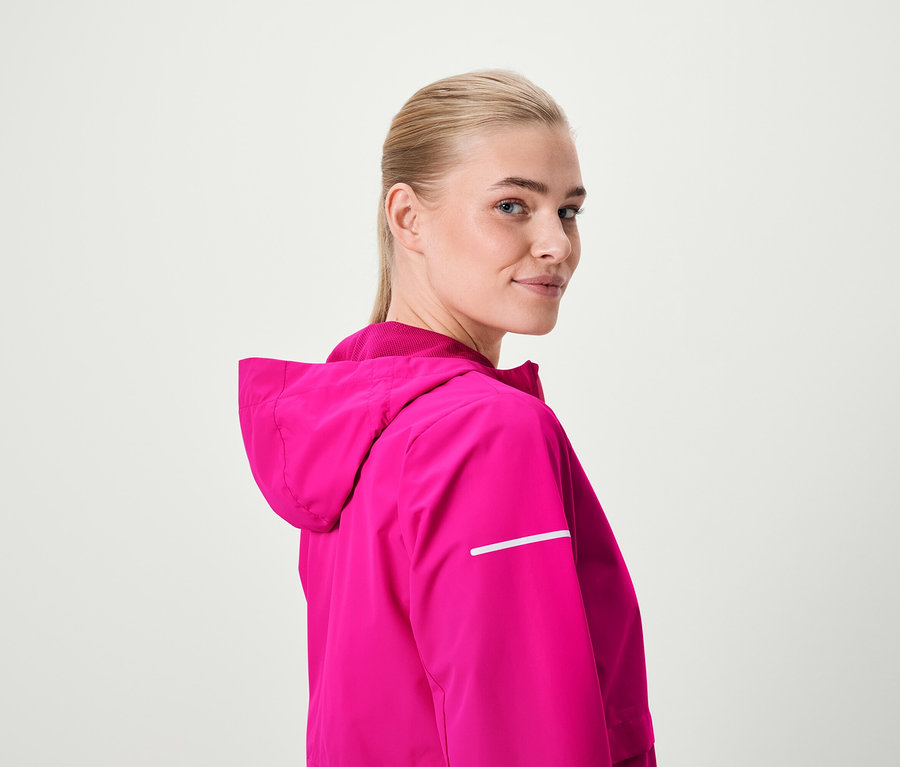 Blondínka sa pozerá cez rameno a má na sebe ružovú bundu s kapucňou.