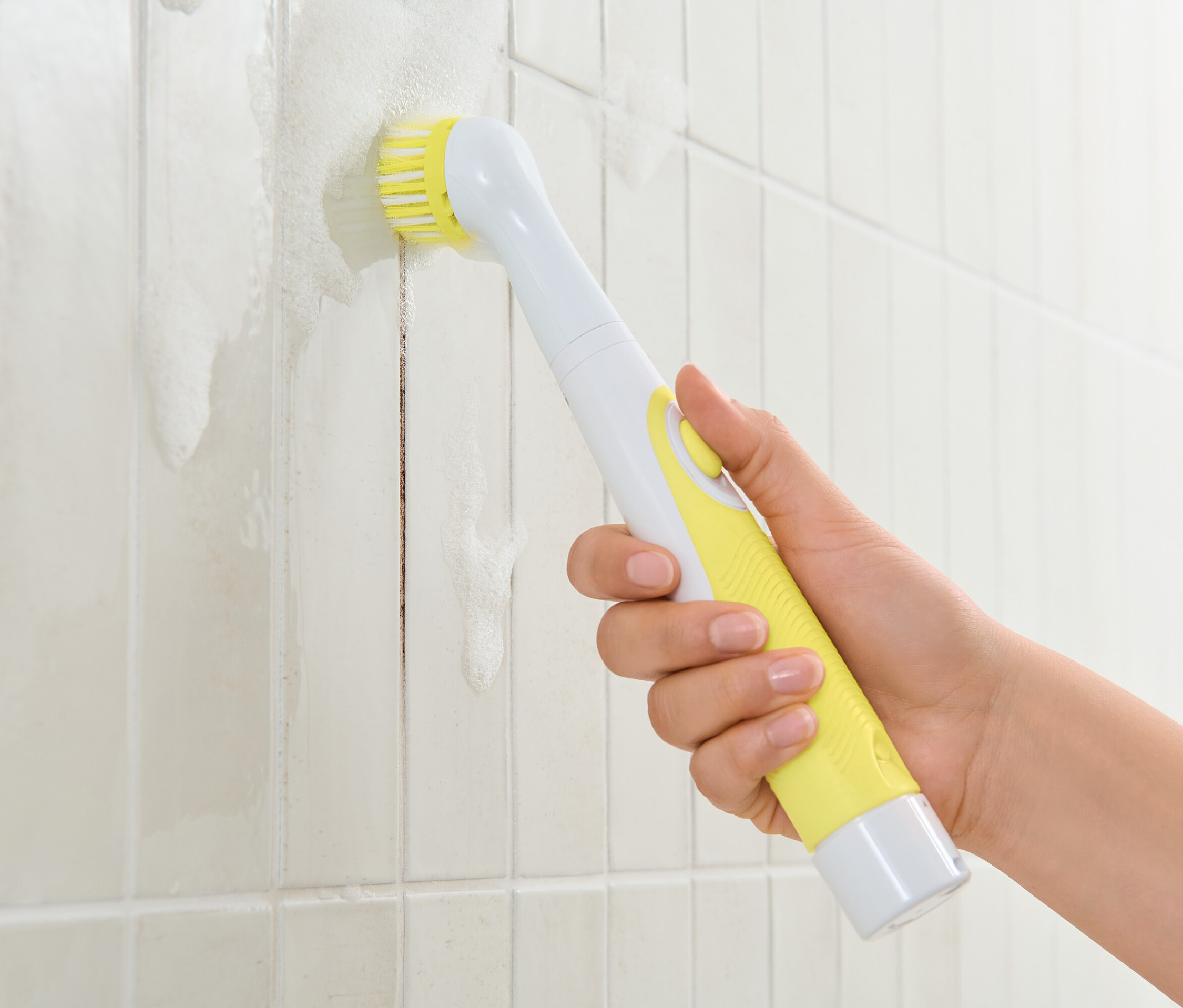 Ruka drží elektrickú čistiacu kefu so žltou rukoväťou a čistí biele dlaždice