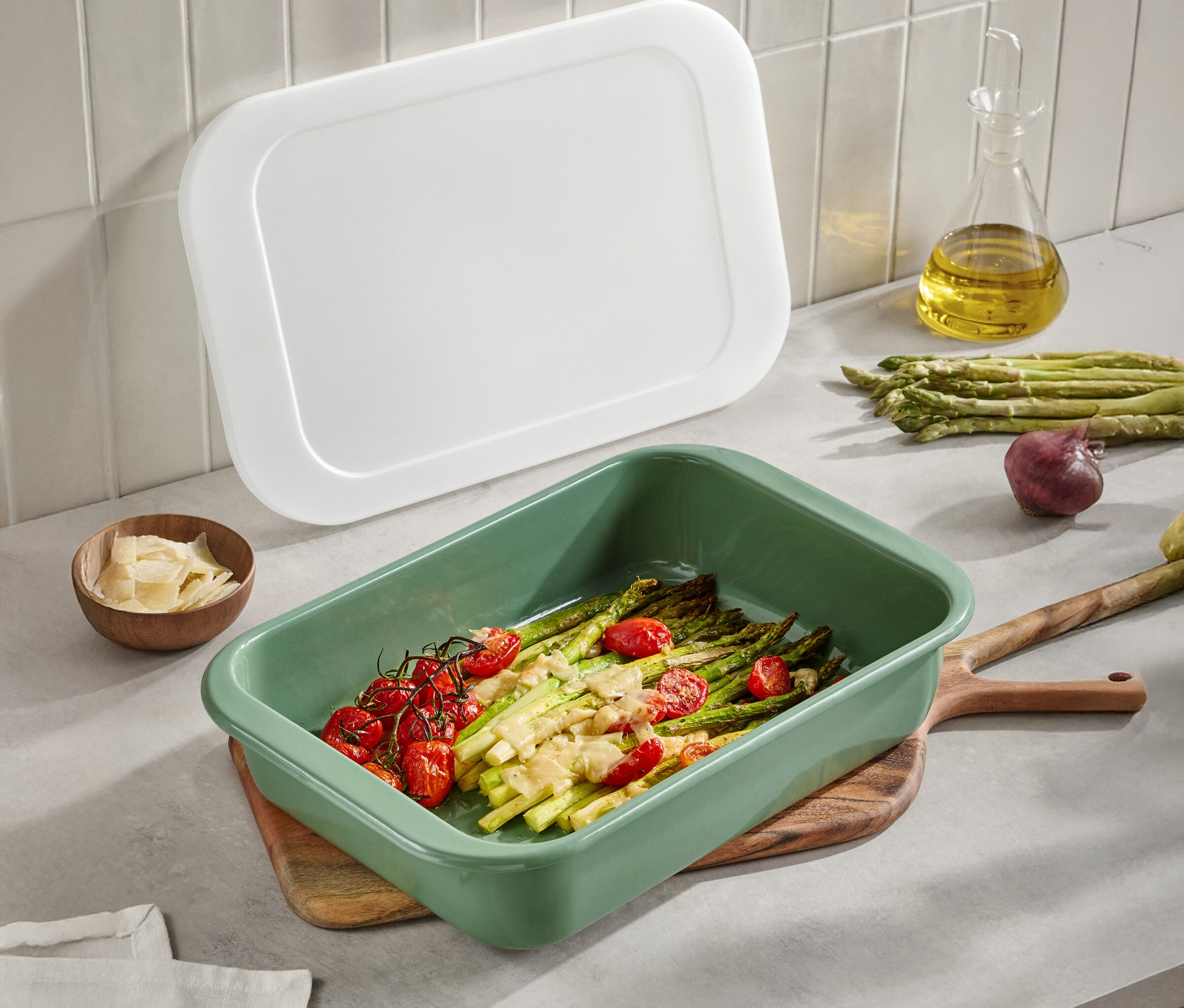 Zelená zapekacia misa so špargľou a paradajkami na drevenom stole. Viditeľné je aj biele veko, olivový olej, parmezán a cibuľa.