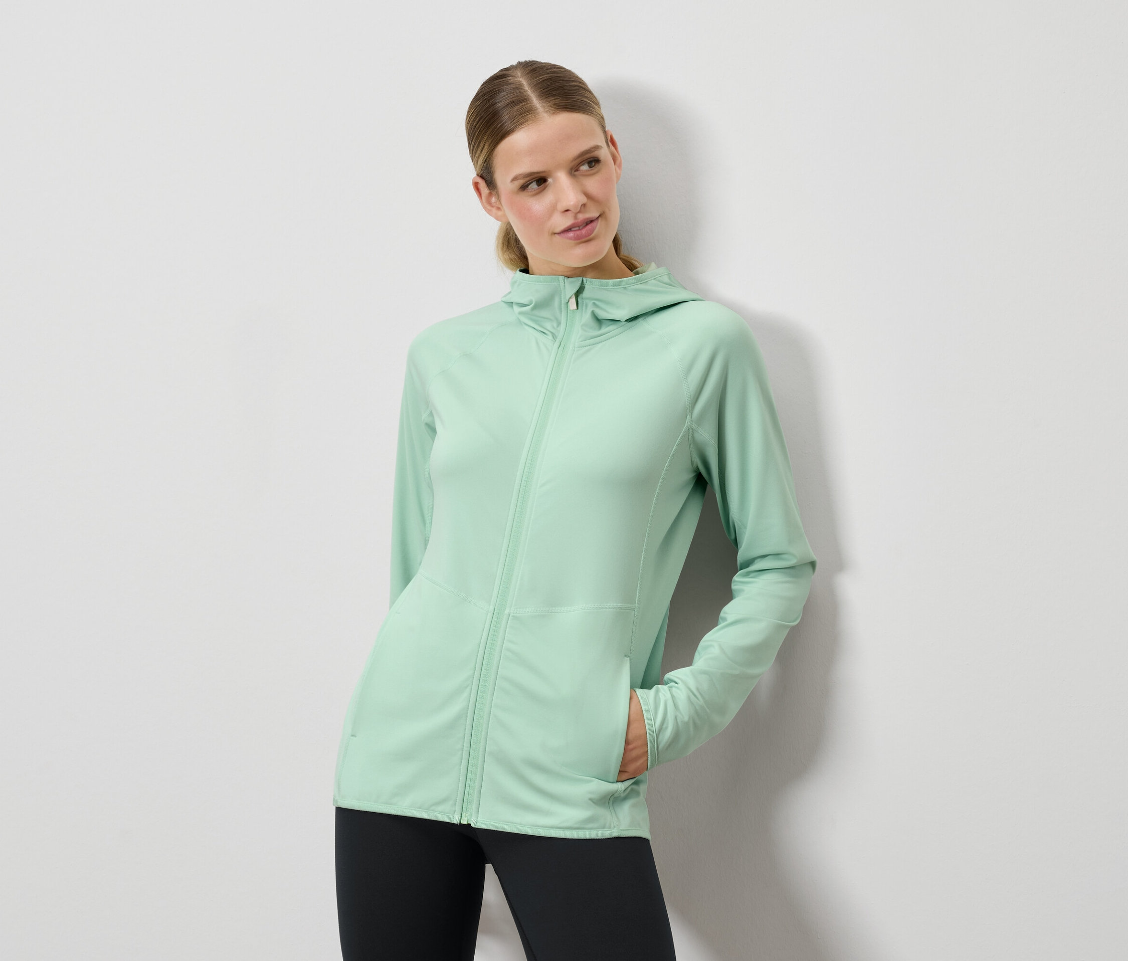 Žena pózuje v mätovo zelenom svetri s kapucňou a čiernych legínach.