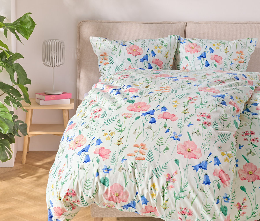 Posteľ s kvetinovou posteľnou bielizňou, vedľa nej stolík s lampou a knihami, vľavo rastlina.