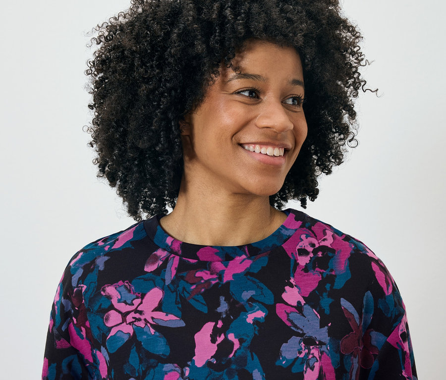 Portrét usmievajúcej sa ženy s afro účesom, oblečenej v čiernom tope s kvetinovým vzorom v ružovej a modrej farbe.