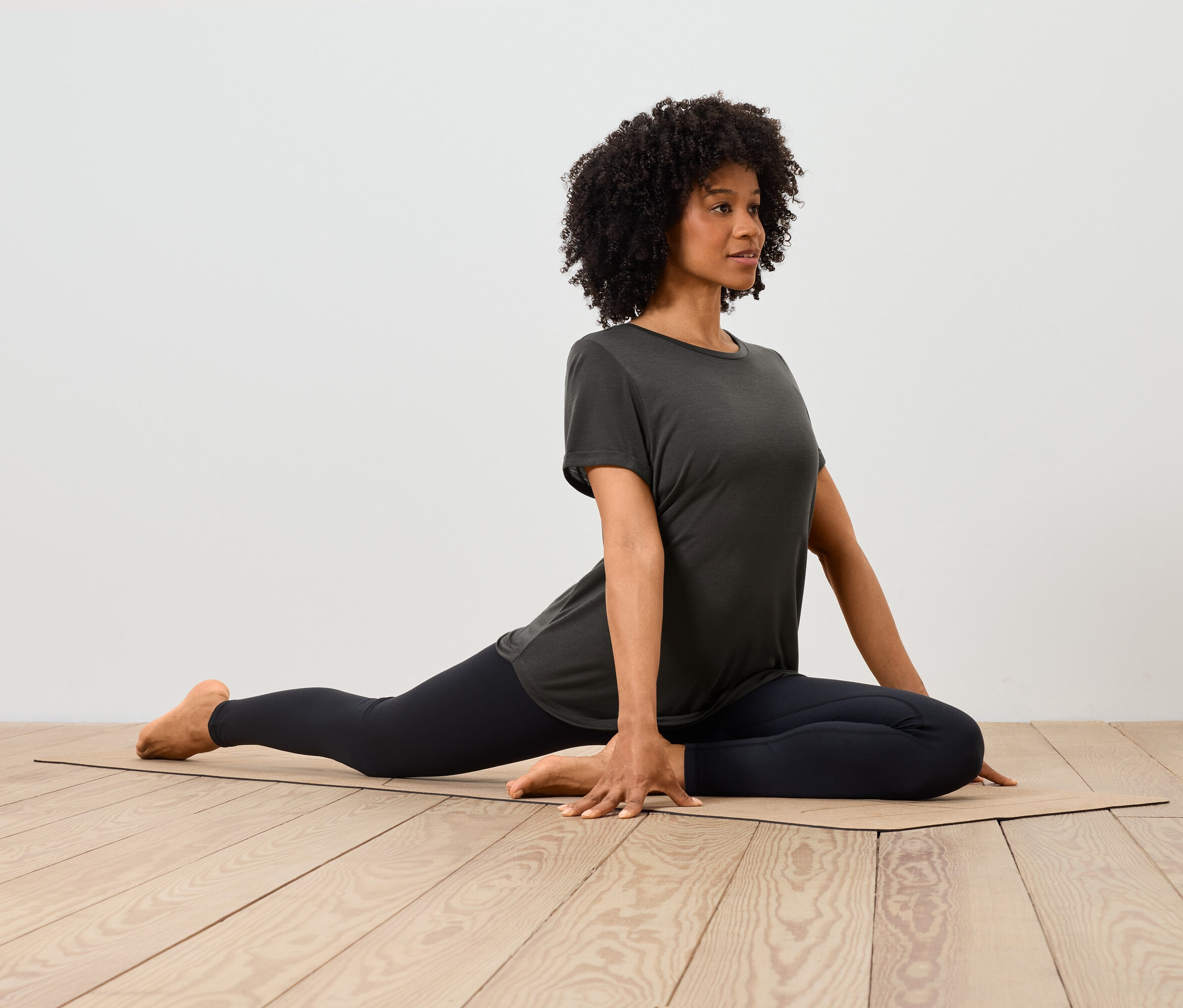Afroamerická žena v tričku a legínach sa naťahuje na podložke na drevenej podlahe.
