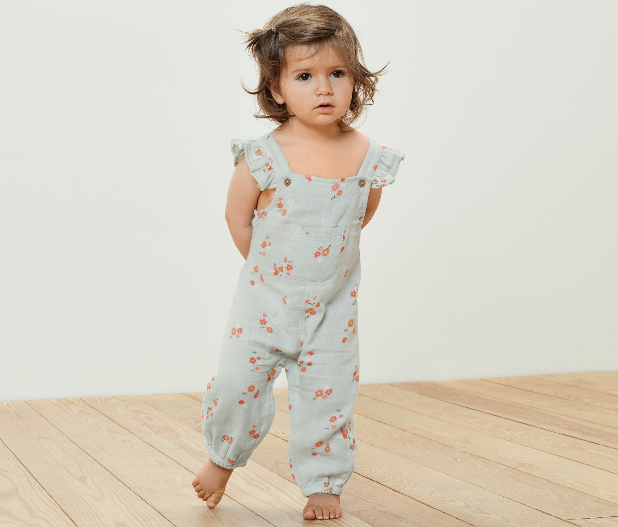 Batoľa stojí na drevenej podlahe a má na sebe detský mušelínový overal s kvetinovým vzorom.