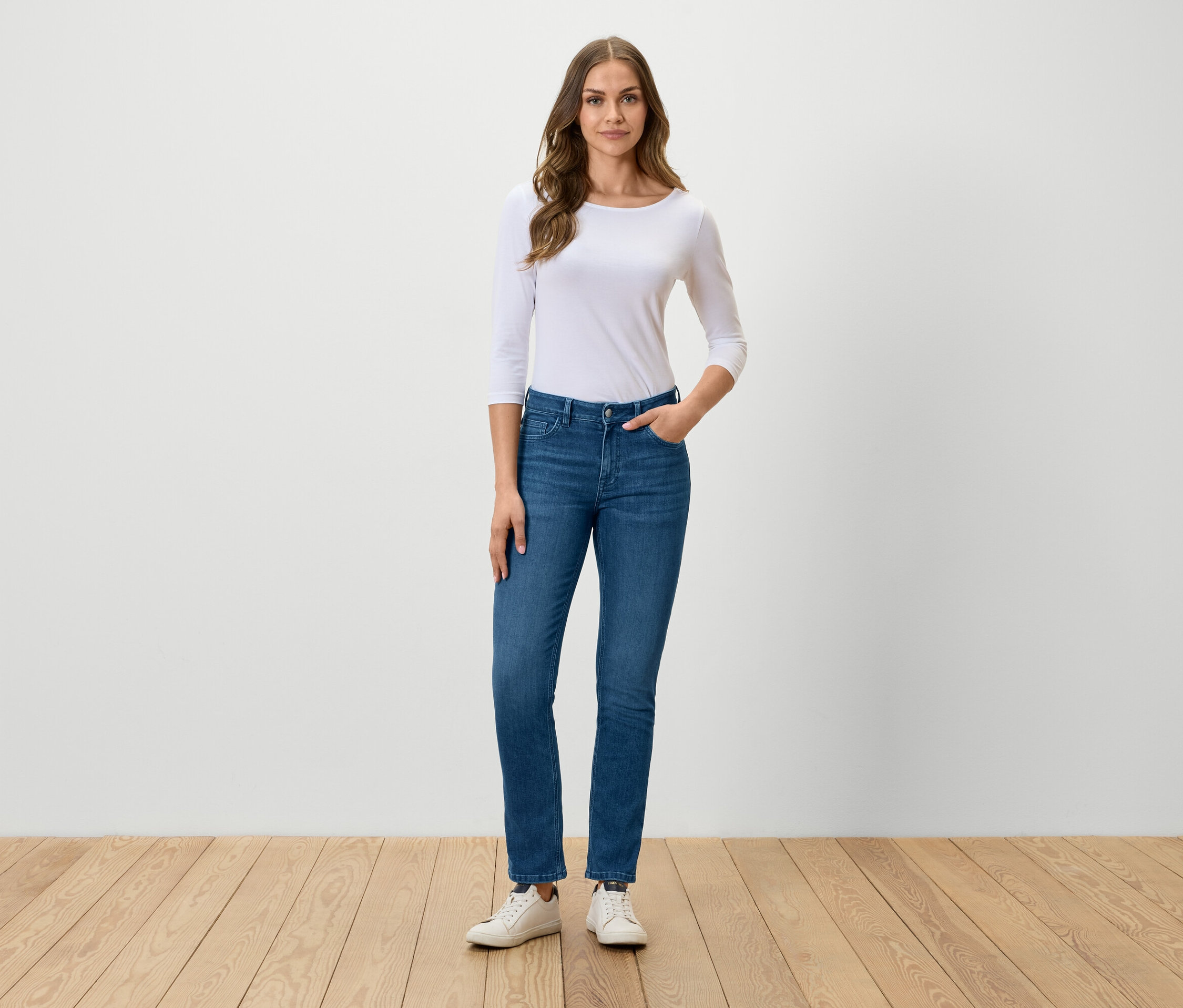 Žena stojí a pózuje v modrých džínsoch, bielom tričku a bielych teniskách.