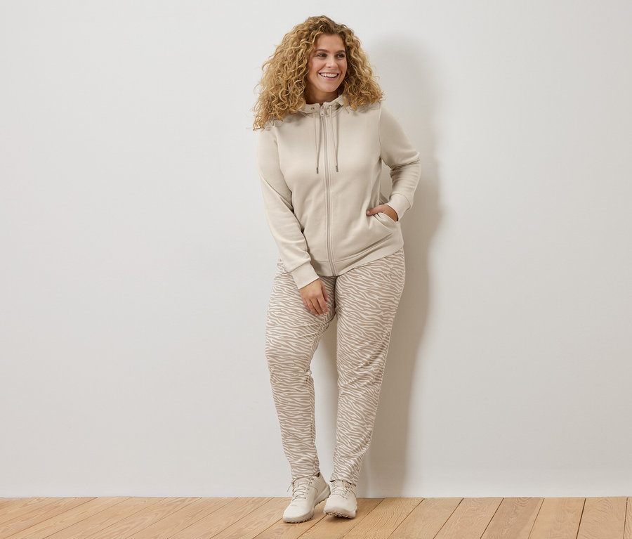 Žena s kučeravými vlasmi v béžovej mikine s kapucňou a nohaviciach so vzorom zebry sa opiera o bielu stenu.