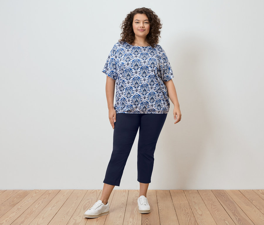 Žena s kučeravými vlasmi stojí pred stenou a má na sebe modrú vzorovanú blúzku, tmavomodré nohavice a biele tenisky.